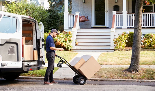 A delivery person using a hand truck to move a large box from a van in front of a house.